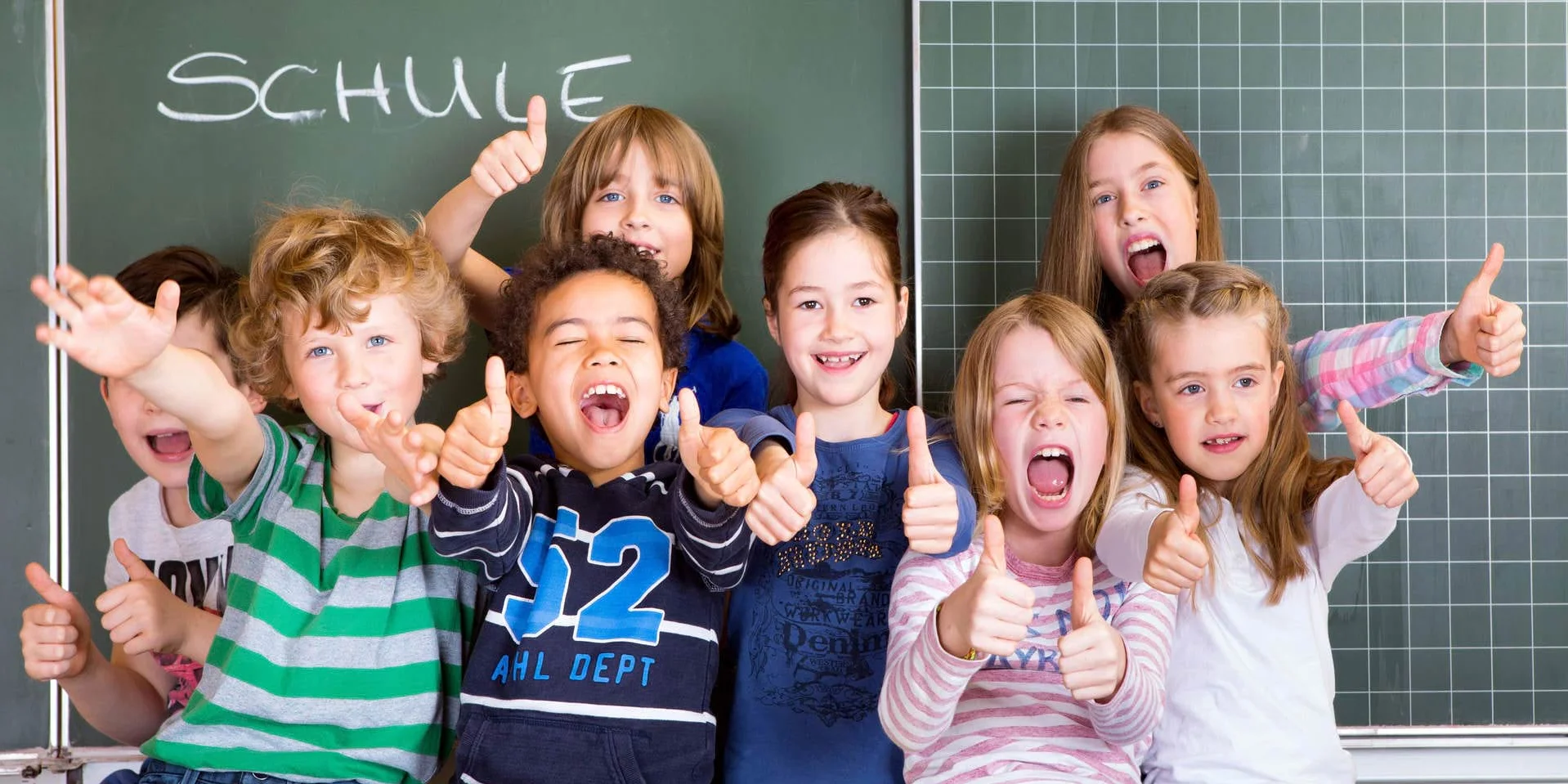 Kinder sitzen vor Tafel und halten Daumen nach oben Kinder sitzen vor Tafel und halten Daumen nach oben